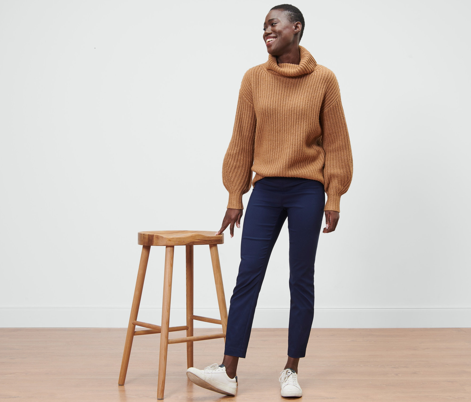 Žena stojí vedle dřevěné stoličky a má na sobě modré strečové kalhoty.