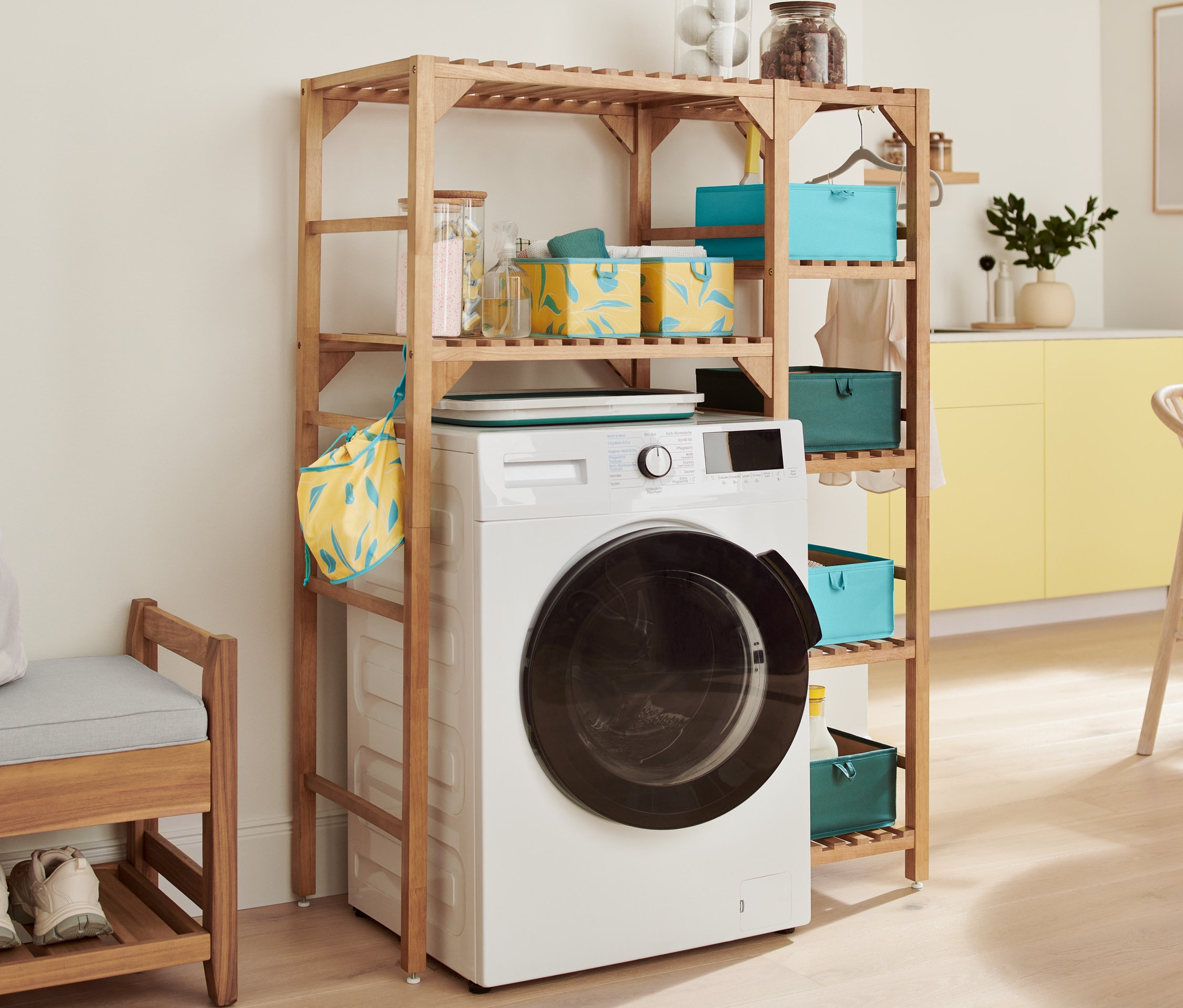 Bílá pračka je umístěna pod dřevěným regálem. Na regále jsou úložné boxy do zásuvky, skládací koš na prádlo a regál na pračku. Vlevo je lavice s úložným prostorem.