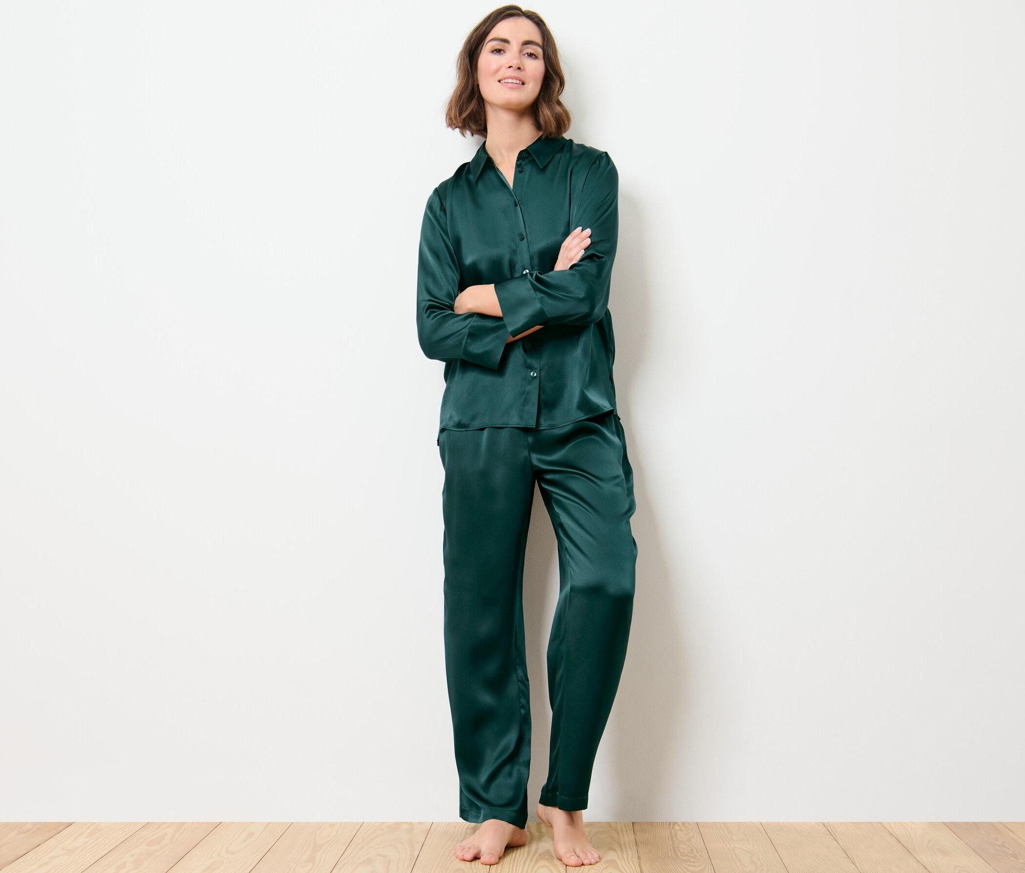 Žena stojí s rukama zkříženýma na prsou a má na sobě tmavě zelené hedvábné pyžamo. Její pyžamový top z čistého hedvábí je zapnutý.