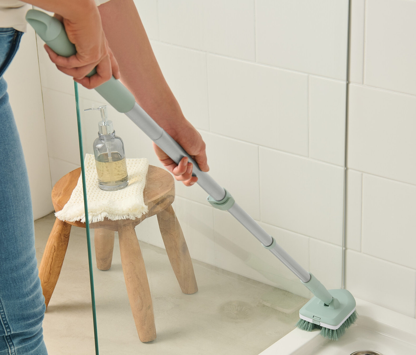 Žena čistí sprchový kout kartáčem na čištění dlaždic a koupelny. Na stoličce stojí dávkovač mýdla a ručník.