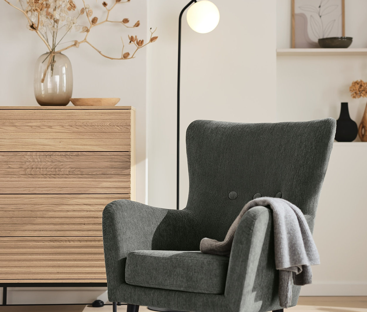 Antracitové polstrované křeslo s přehozenou dekou stojí vedle komody »Finnsbo« a černé stojací lampy se skleněnou koulí.