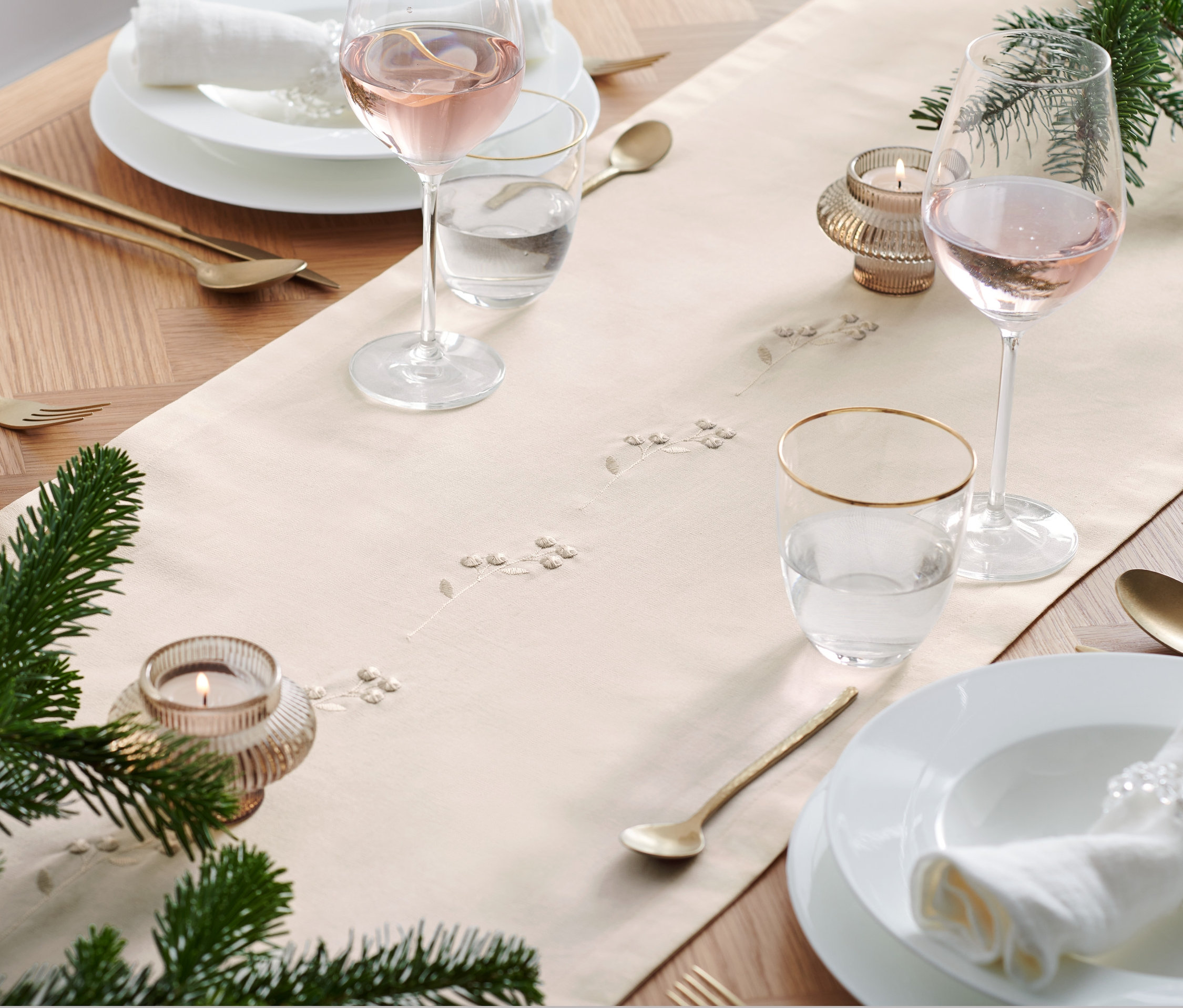 Prostřený stůl s vyšívaným běhounem, sklenicemi, talíři a zlatými příbory.