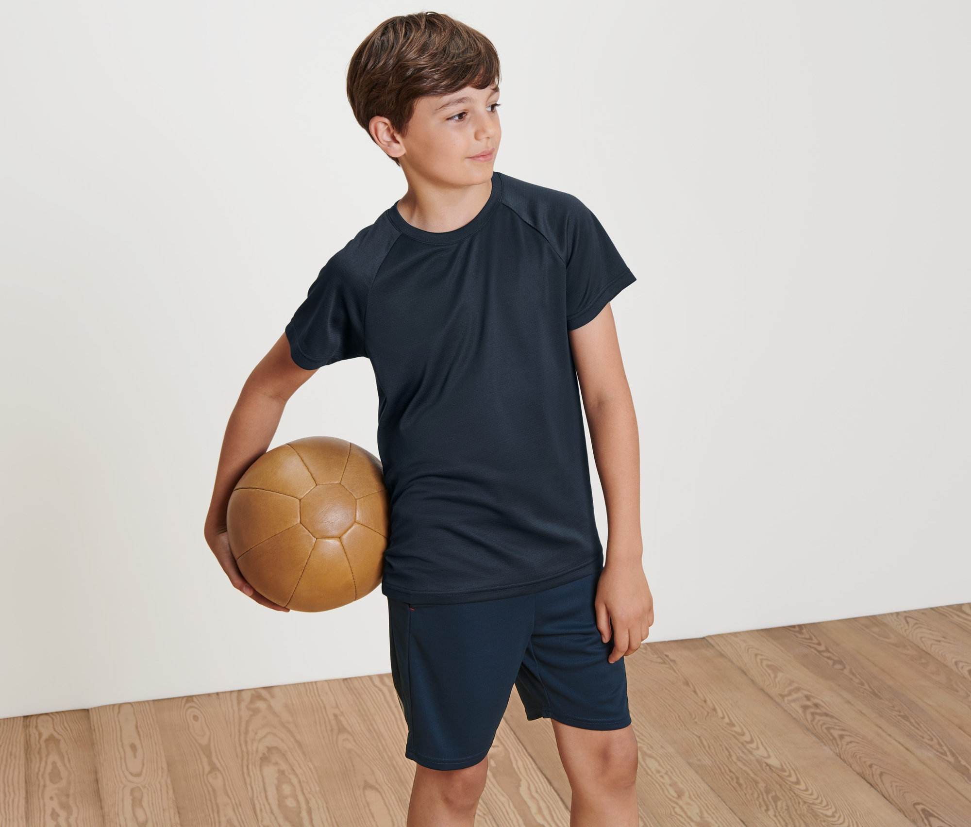 Chlapec drží hnědý kožený míč. Má na sobě jedno ze dvou dětských funkčních triček.