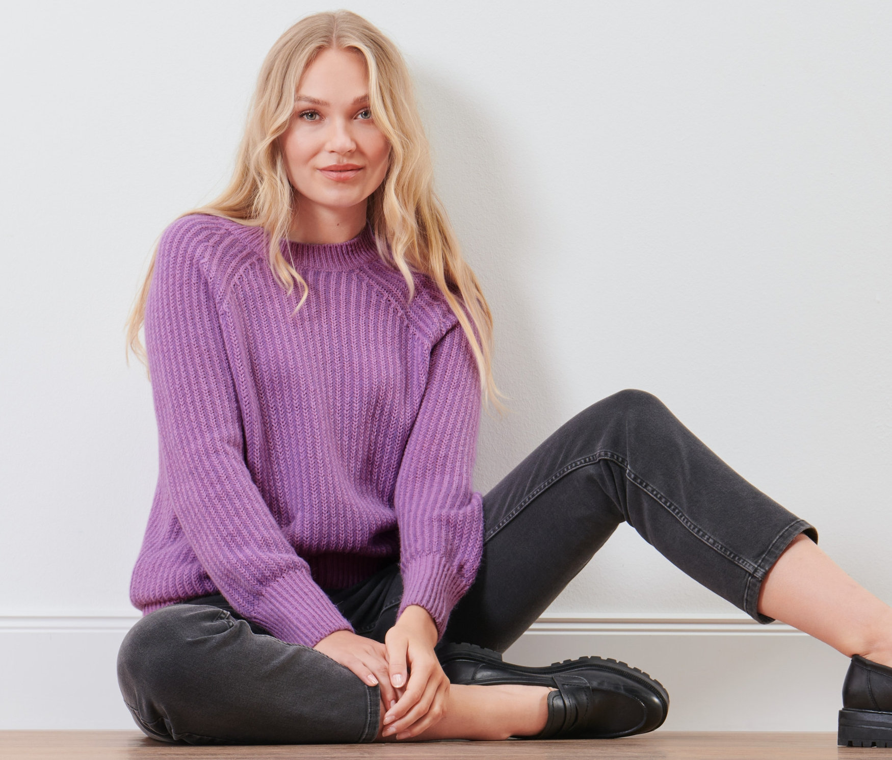 Blonďatá žena sedí na podlaze a má na sobě šeříkový pletený svetr.