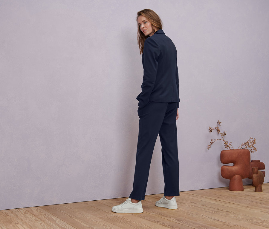 Žena se dívá přes rameno a má na sobě tmavě modré žerzejové sako, tmavě modré žerzejové kalhoty s puky a bílé triko s polodlouhým rukávem.