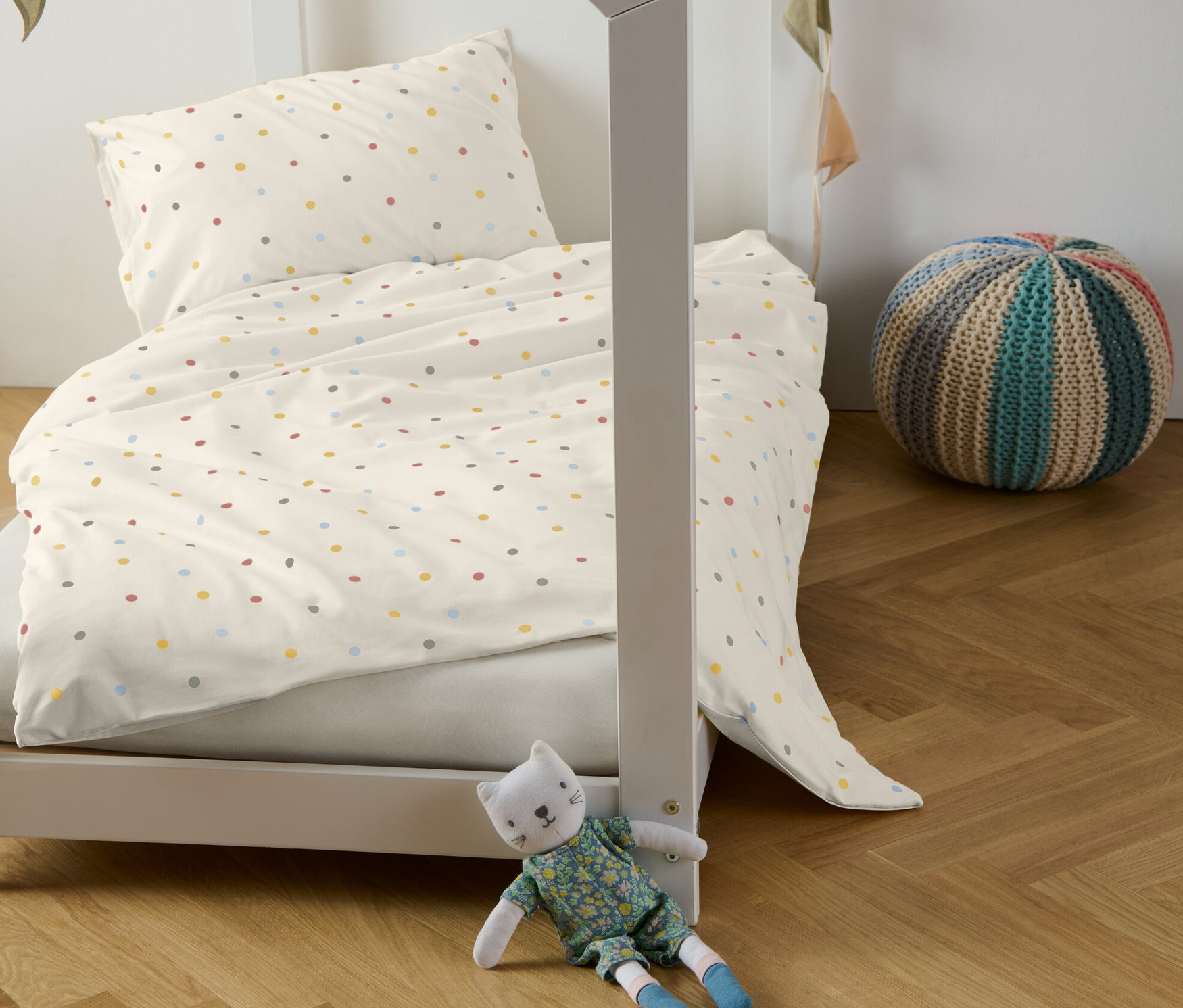 Dětská postel s povlečením renforcé s barevnými puntíky, dekorativní girlanda a plyšová kočka.