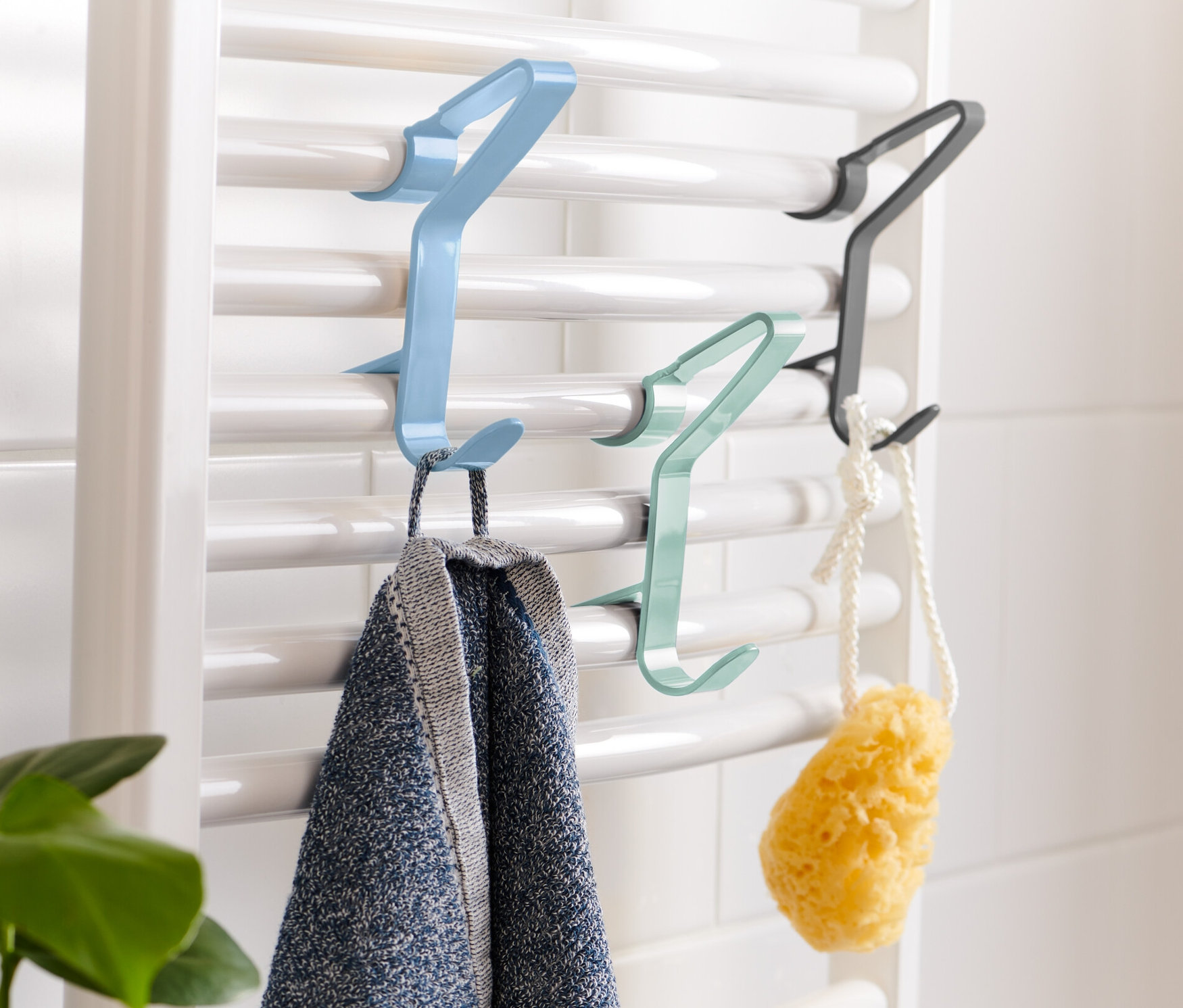 Na bílém topení visí modrý, zelený a černý háček na topná tělesa. Na modrém háčku visí ručník.