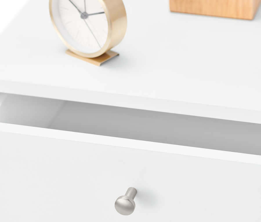 Detail bílého nočního stolku s otevřenou zásuvkou, zlatými hodinami a dřevěnou krabičkou.