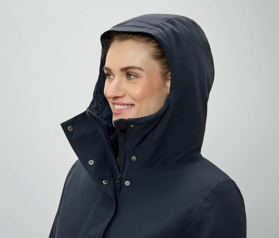 Žena s kapucí na hlavě a tmavě modré bundě se dívá doprava.