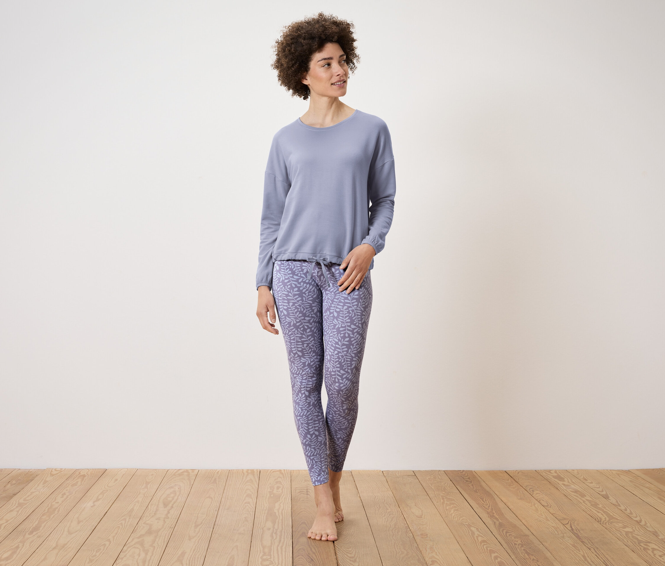 Žena v světle modrém pyžamu s potiskem listů stojí na dřevěné podlaze.