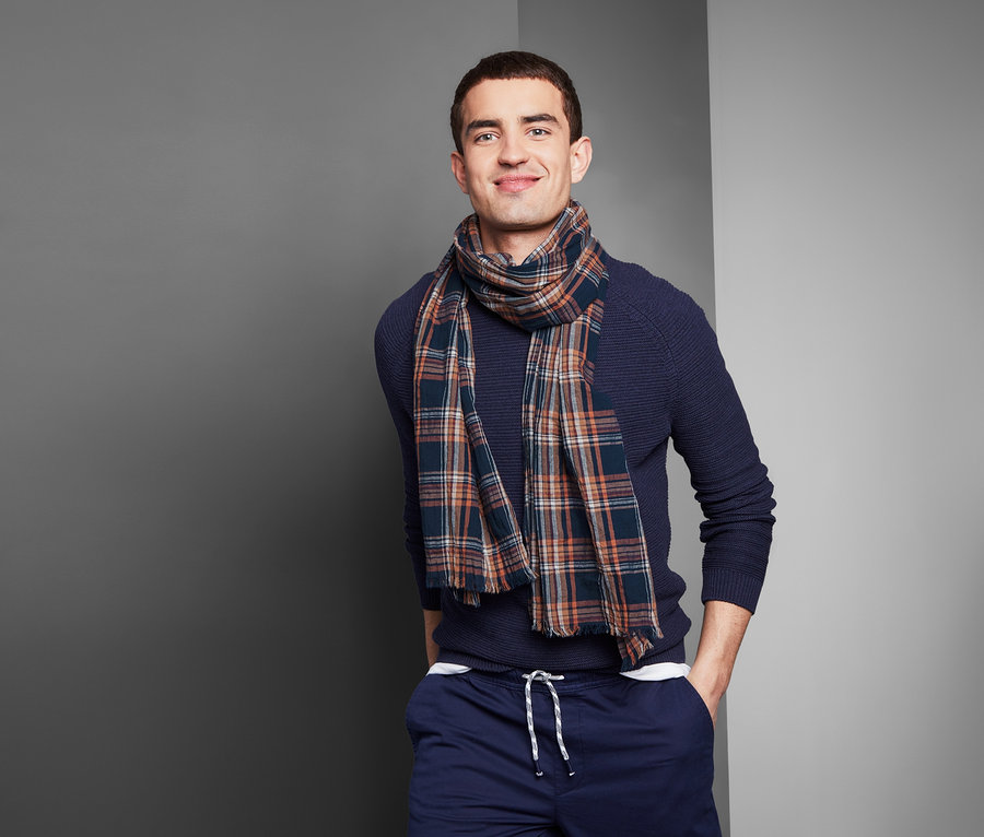 Muž s úsměvem pózuje v modrém svetru a kostkované tkané šále.