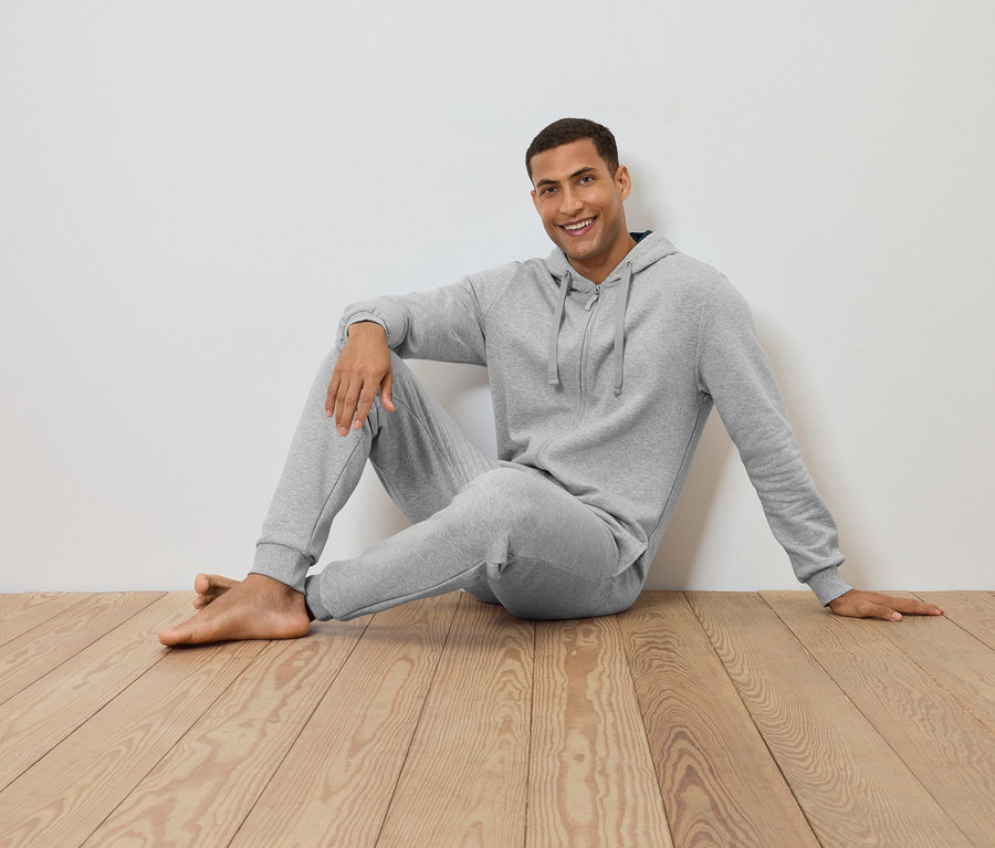 Muž sedí na dřevěné podlaze opřený o bílou zeď a má na sobě šedou mikinu s kapucí a tepláky.