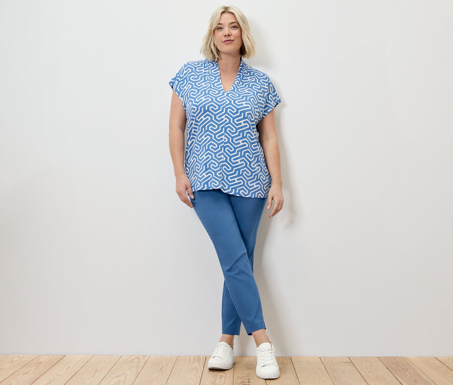 Blonďatá žena pózuje ve strečových kalhotách a halence s výstřihem do V, obojí modré.