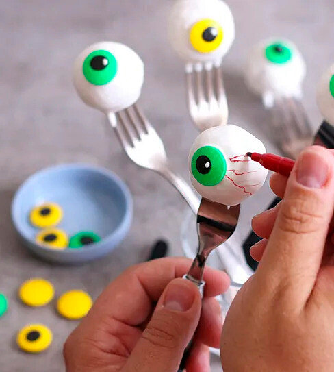 Ruka kreslí červené žilky na bílou oční bulvu s duhovkou na vidličce pomocí červeného fixu.