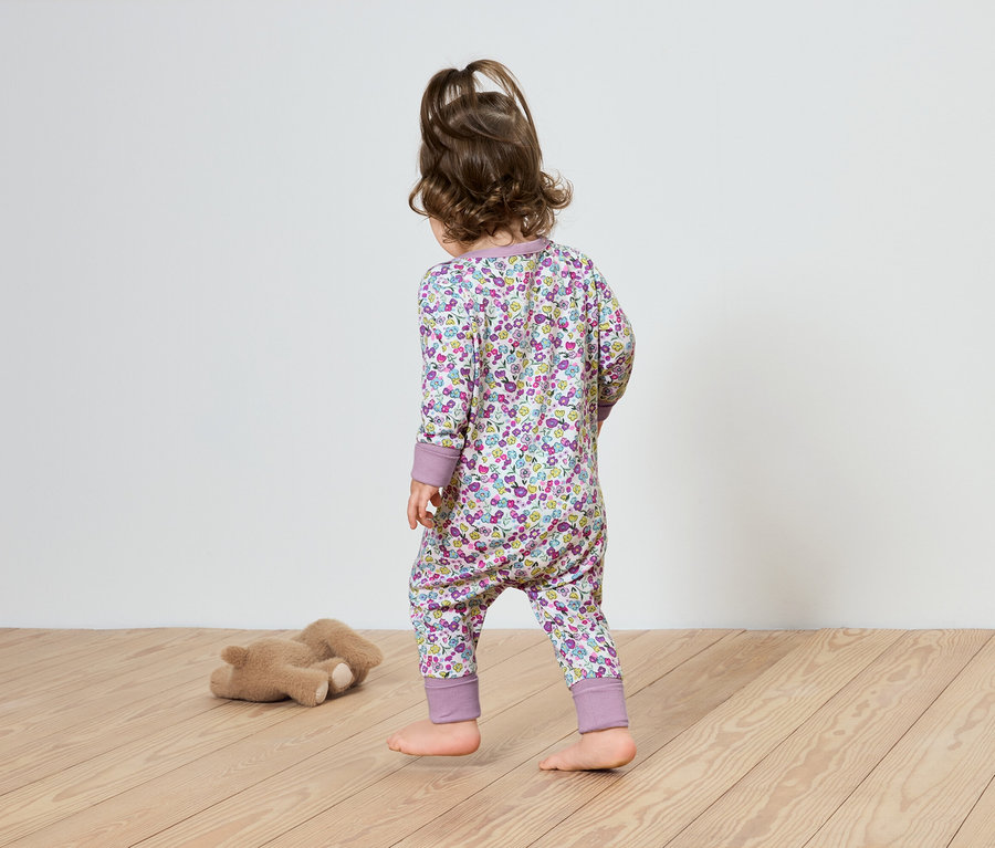 Dítě v květinovém overalu kráčí po dřevěné podlaze směrem k plyšovému medvídkovi.