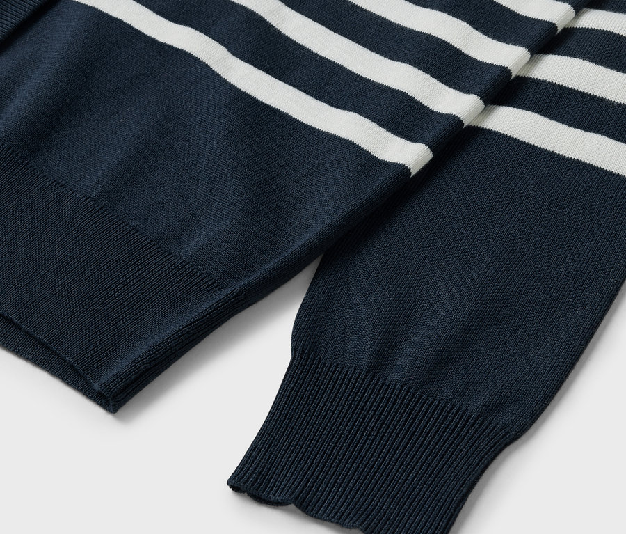 Detail modrého svetru s bílými pruhy, viditelné rukávy a žebrovaný lem.