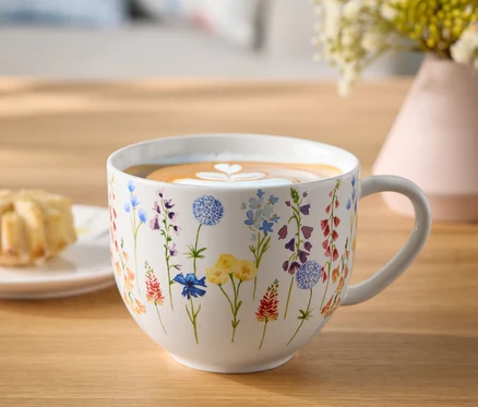 Květinový hrnek s kávou a latte art srdce na dřevěném stole vedle talíře s pečivem a vázy s květinami.