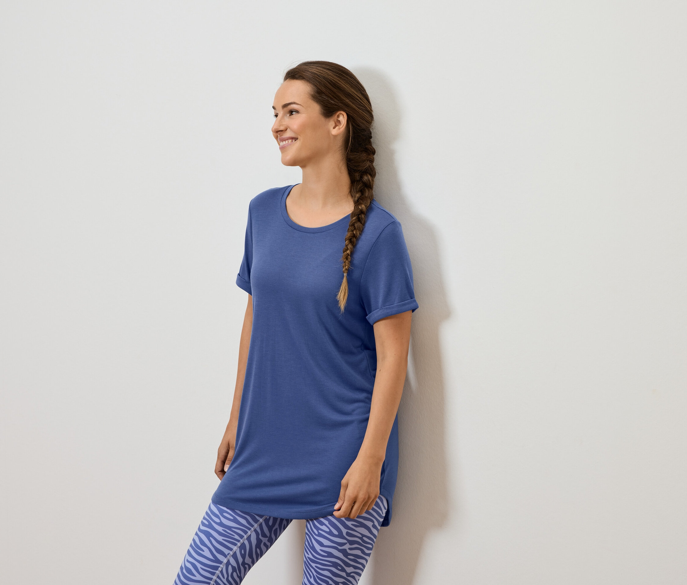 Žena s hnědým copem stojí u bílé zdi, má na sobě modré tričko a modré legíny se zvířecím vzorem.