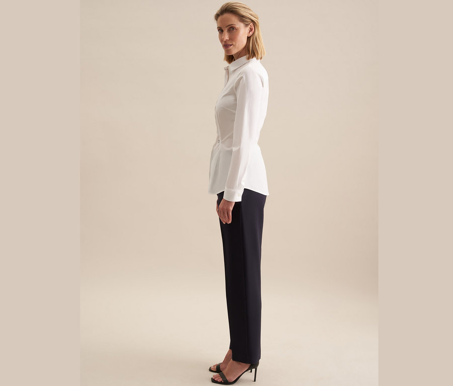 Žena stojící bokem má na sobě bílou halenku slim fit Seidensticker a tmavě modré kalhoty.
