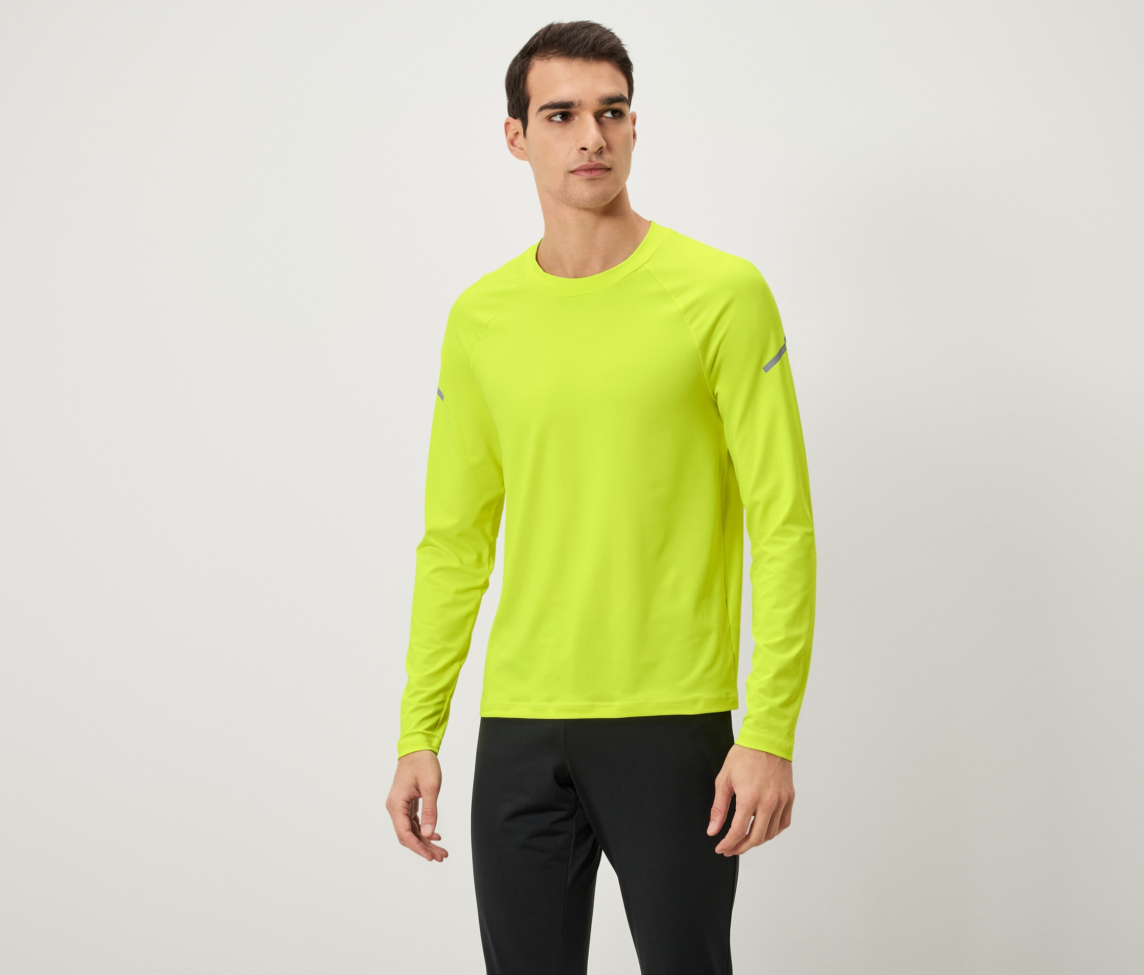 Muž pózuje v neonově žlutém tričku s dlouhým rukávem a černých teplácích.