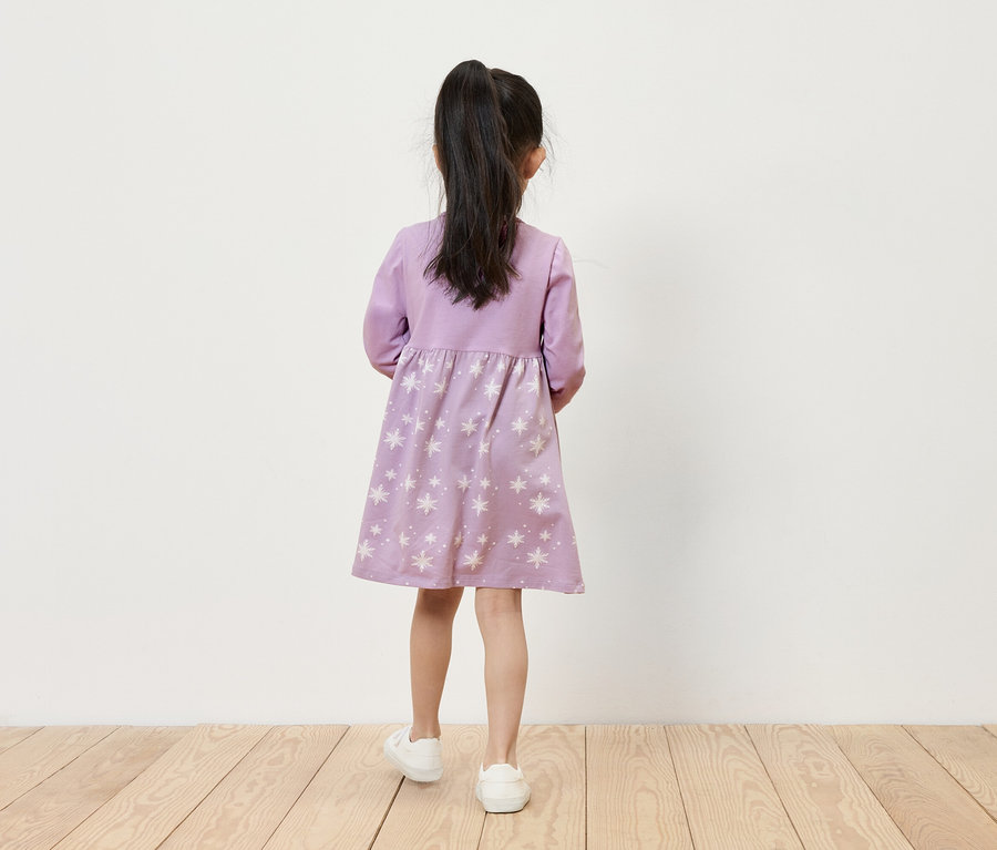 Dívka v lila šatech s hvězdami a bílých teniskách stojí zády k fotoaparátu.