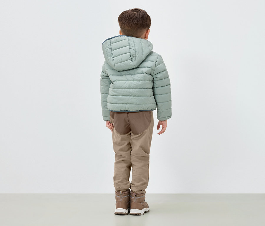 Chlapec stojí zády, má na sobě světle zelenou péřovou bundu s kapucí, hnědé kalhoty a hnědé boty.