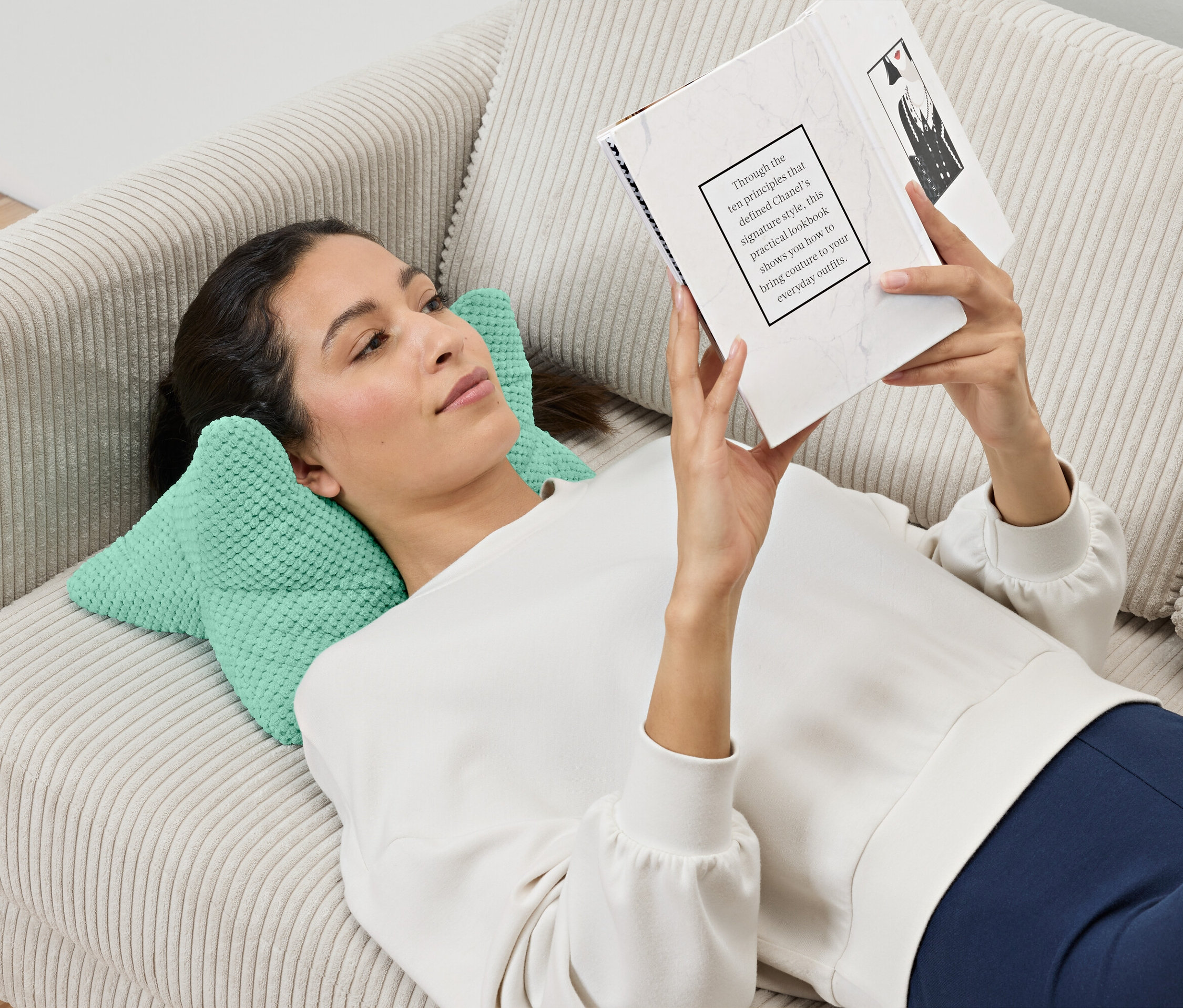 Žena leží na pohovce a čte si knihu, hlavu má položenou na polštáři na čtení.