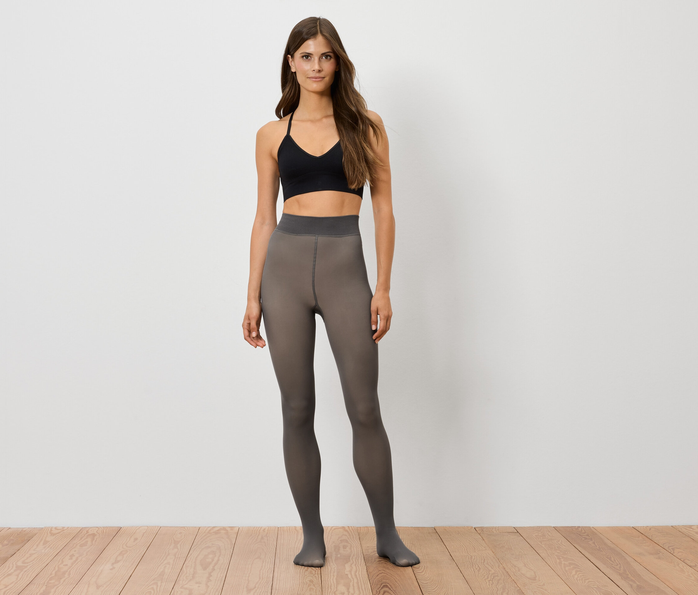 Žena stojí a má na sobě černé sportovní tílko a šedé punčochové kalhoty.