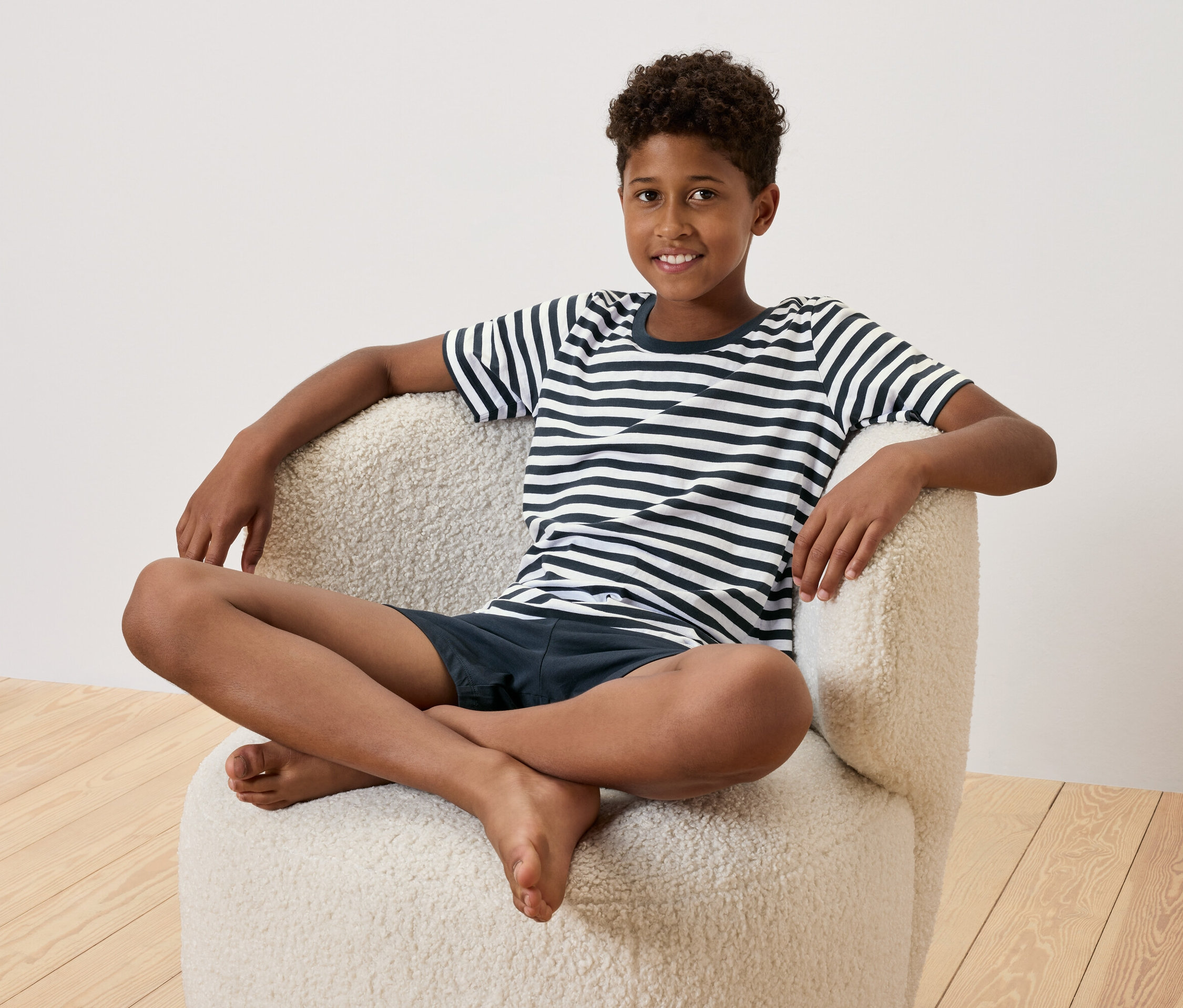 Mladý chlapec sedí s zkříženýma nohama v krémovém křesle. Má na sobě pruhované tričko a tmavě modré šortky.