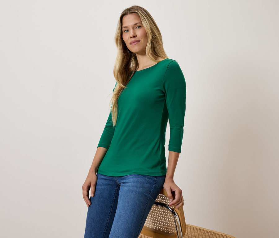 Blondýna v zeleném tričku s tříčtvrtečním rukávem a modrých džínách sedí na židli.