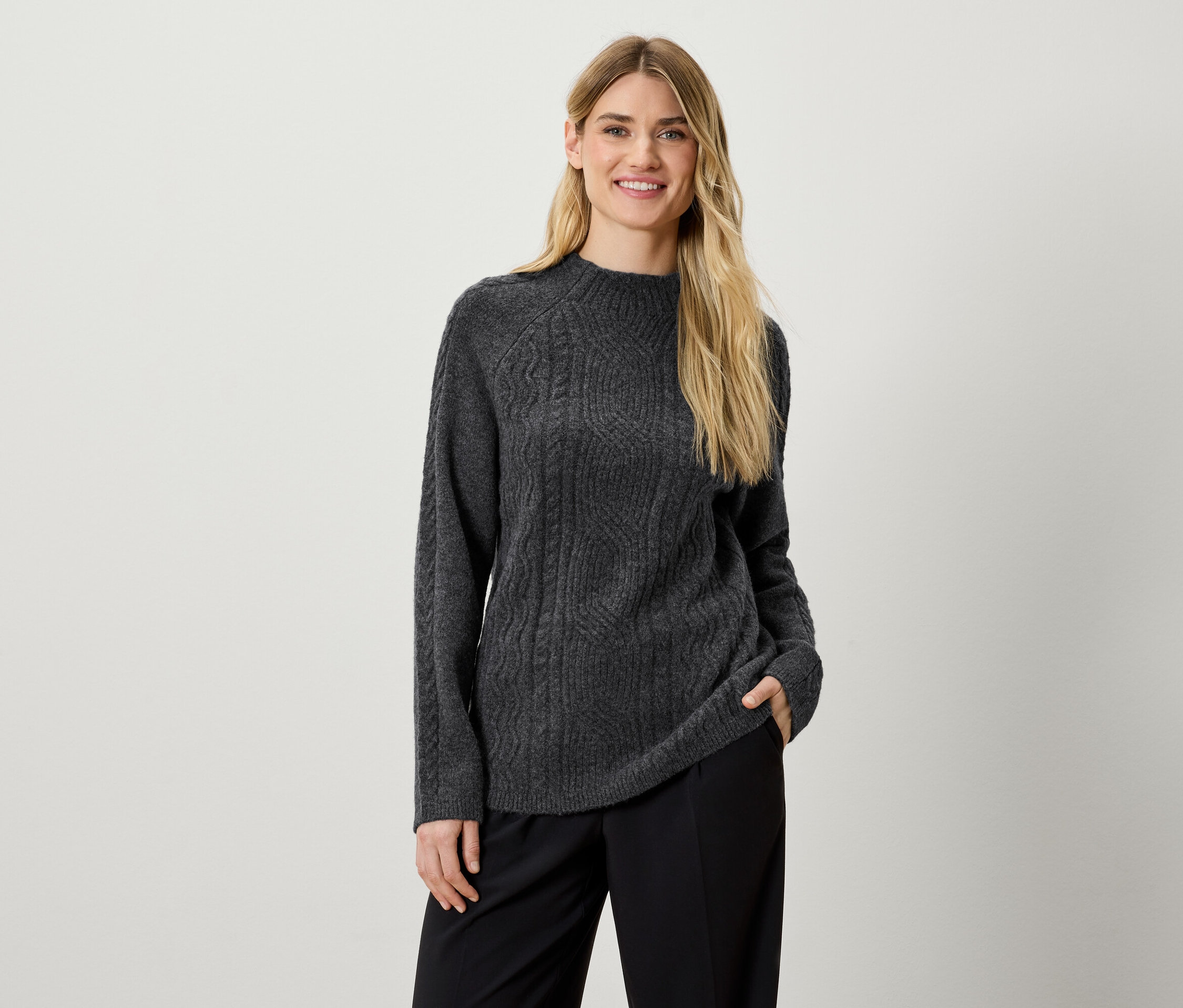 Žena pózuje v šedém pleteném svetru a černých kalhotách.