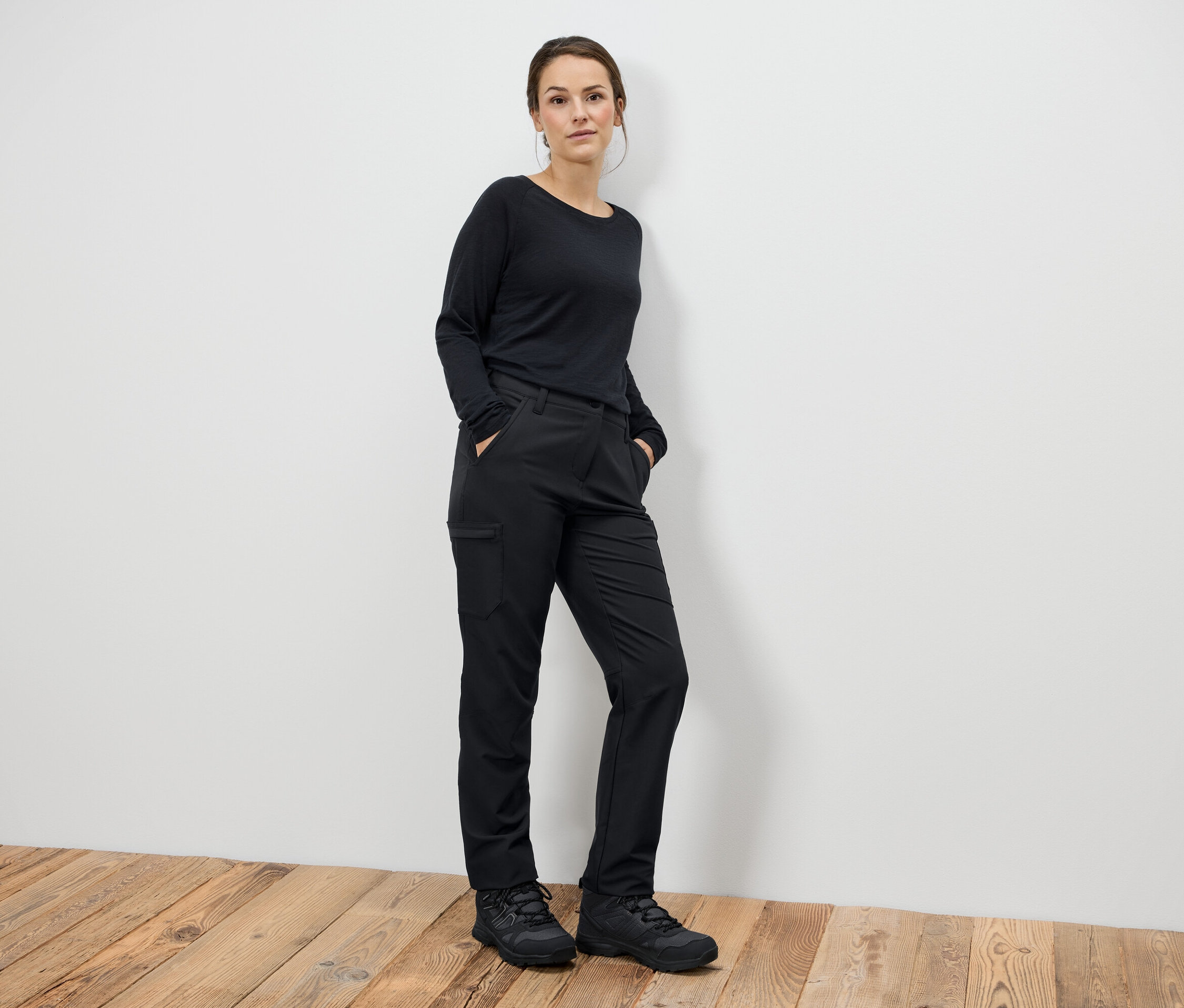 Žena stojí a má ruce v kapsách černých kalhot a černé boty.