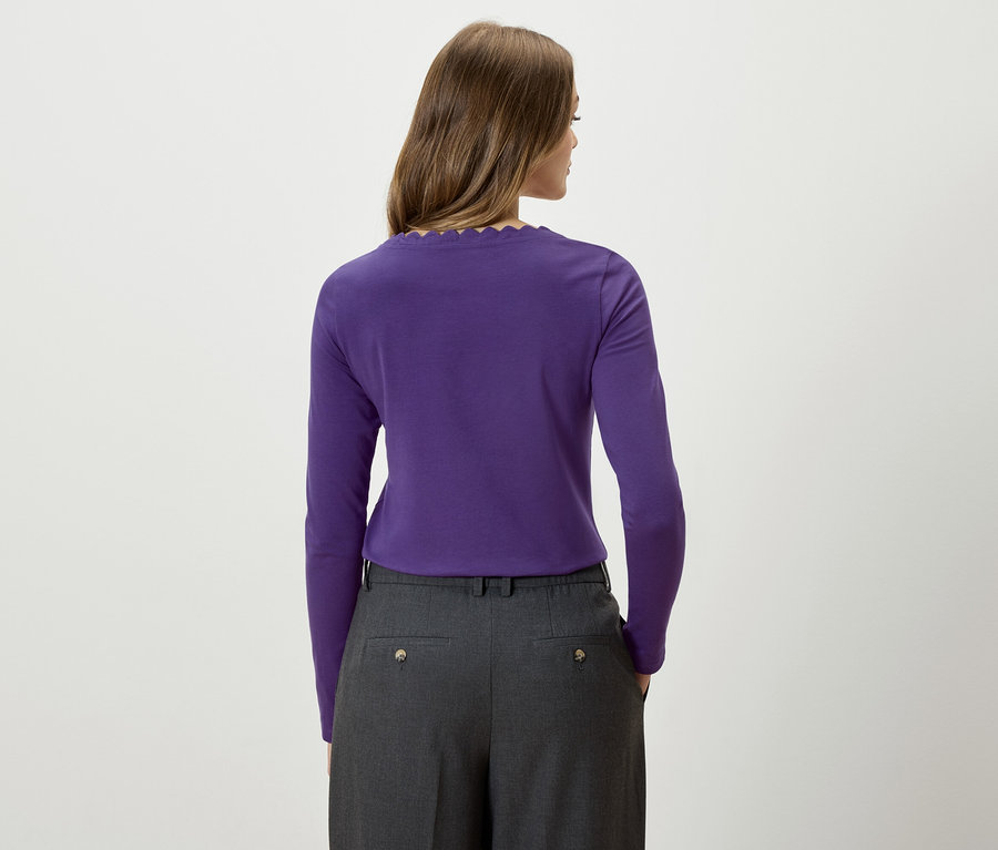 Žena zády k fotoaparátu má na sobě fialové tričko s dlouhým rukávem a šedé kalhoty.