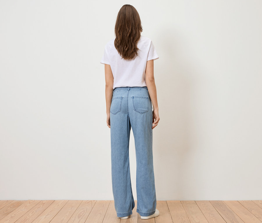 Pohled zezadu na ženu s dlouhými hnědými vlasy, oblečenou v bílém tričku a světle modrých džínách.