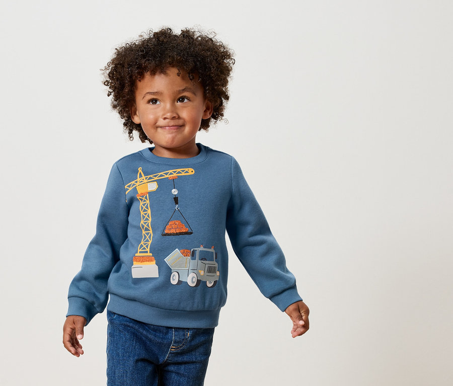 Chlapec s kudrnatými vlasy má na sobě modrou mikinu s aplikací žlutého jeřábu a nákladního auta.