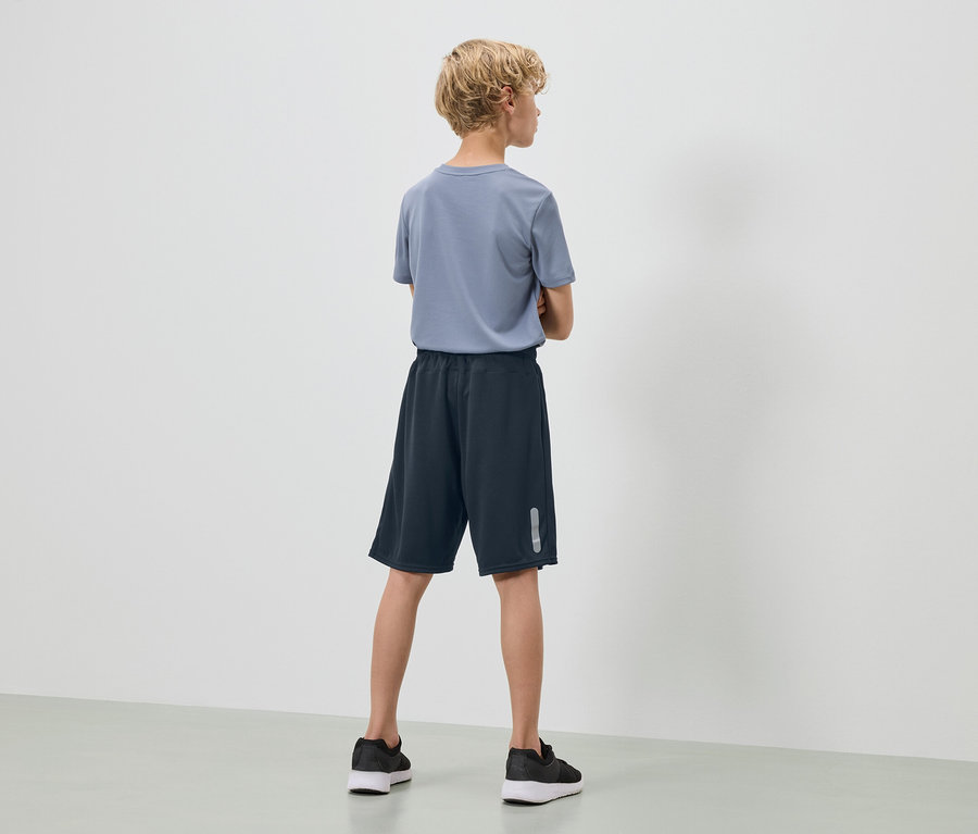 Dospívající s blond kudrnatými vlasy stojí zády k fotoaparátu, má na sobě modré tričko, tmavě modré šortky a černé tenisky.