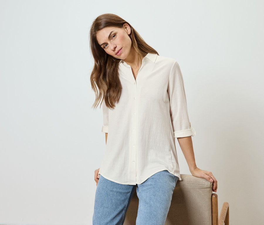 Žena s vlnitými vlasy, oblečená ve světlé košili a džínách, se opírá o židli.