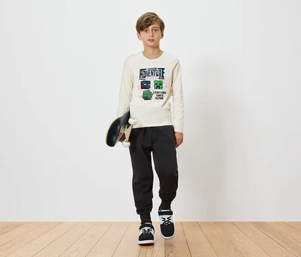 Chlapec drží skateboard a má na sobě světlé tričko s dlouhým rukávem s motivem Minecraftu, kombinované s tmavými tepláky a teniskami na jednoduchém pozadí.