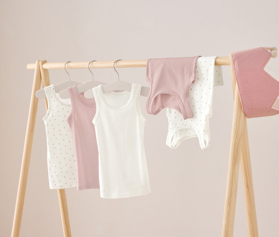 Na dřevěném stojanu visí tři tílka v bílé, růžové a bílé barvě s puntíky, dále růžová košile a bílé body s puntíky. Vpravo visí růžová koruna.