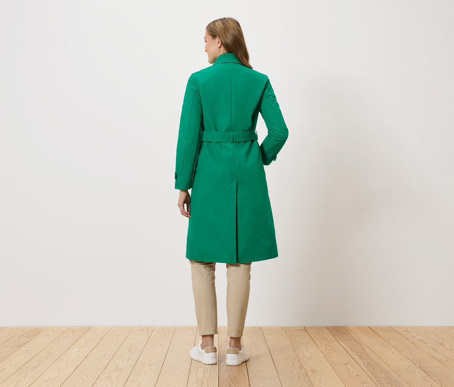 Žena stojí zády ke kameře a má na sobě zelený kabát s páskem, béžové kalhoty a bílé tenisky.