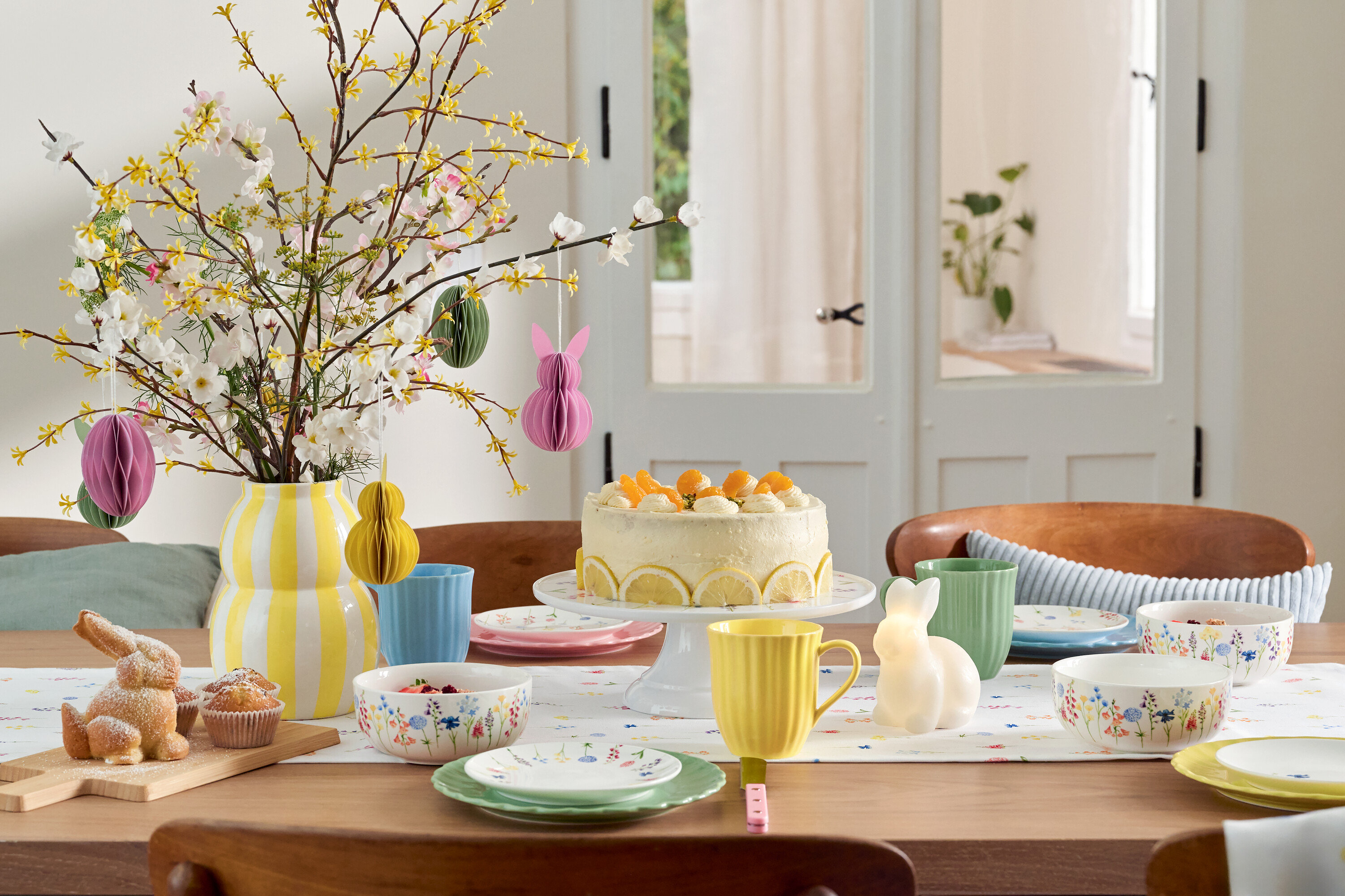 Jídelní stůl s velikonoční výzdobou, dortem, nádobím a vázou s květinami.