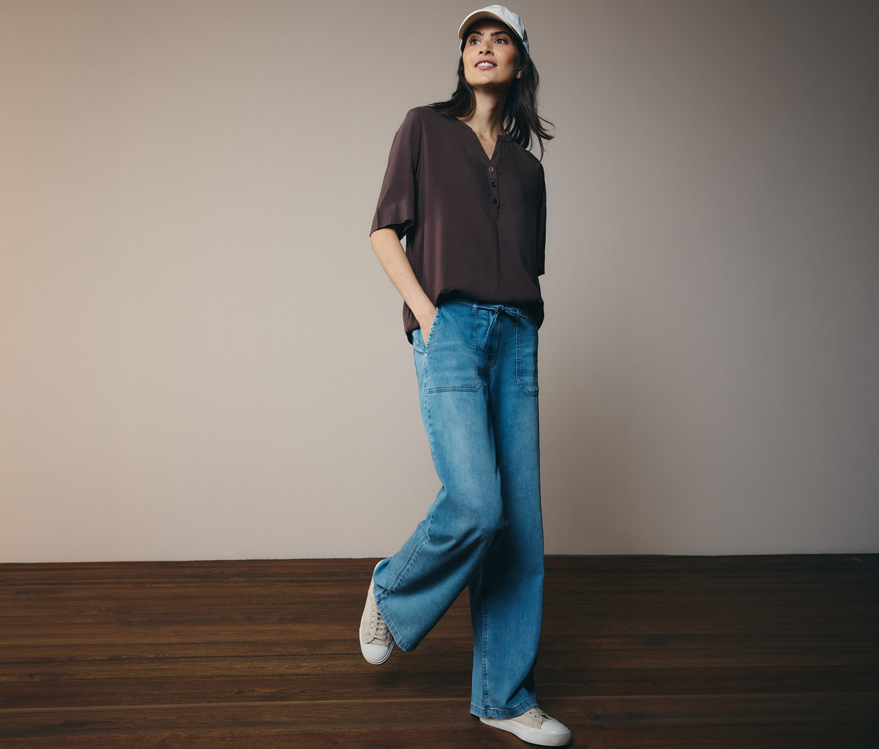 Žena v kšiltovce a hnědé tunikové halence CECIL, džínách a teniskách.