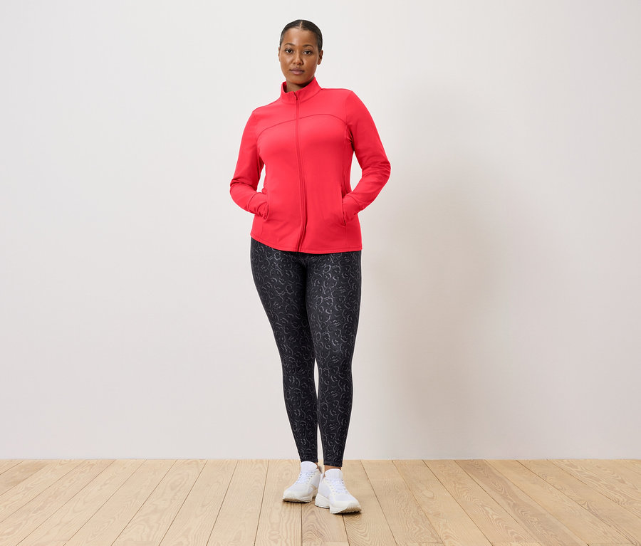 Žena v červené bundě a vzorovaných legínách pózuje před zdí.