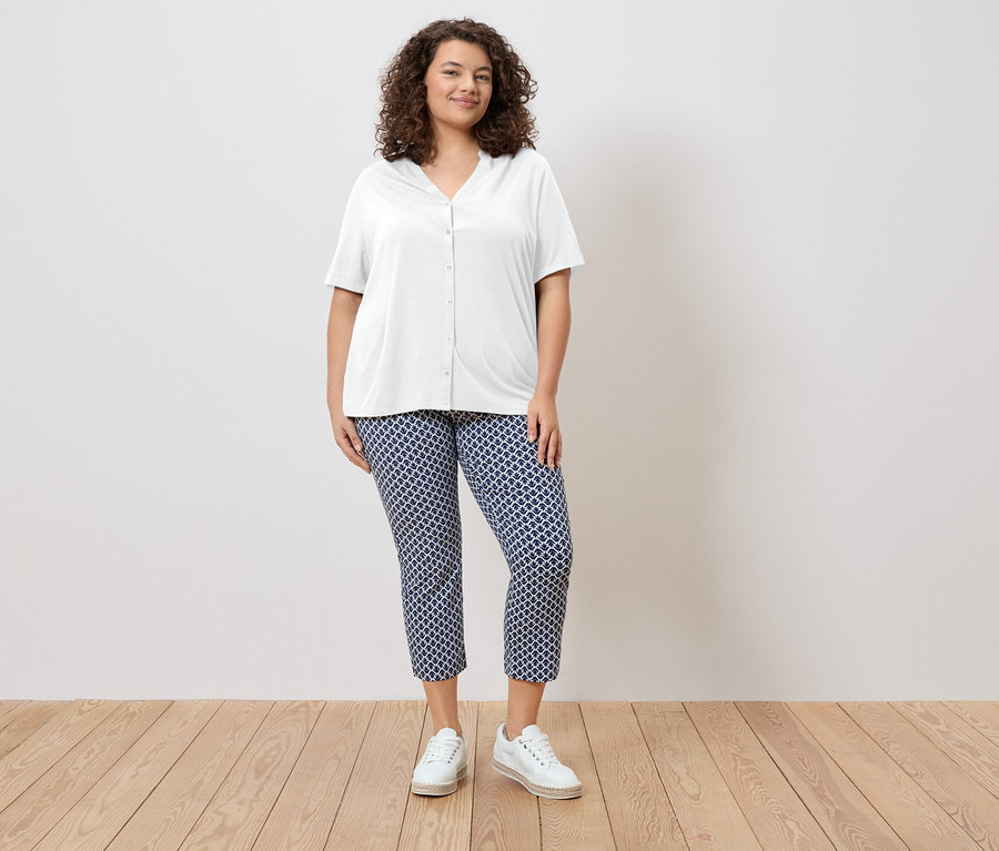 Žena s kudrnatými vlasy má na sobě bílou košili a modré vzorované kalhoty a bílé boty.