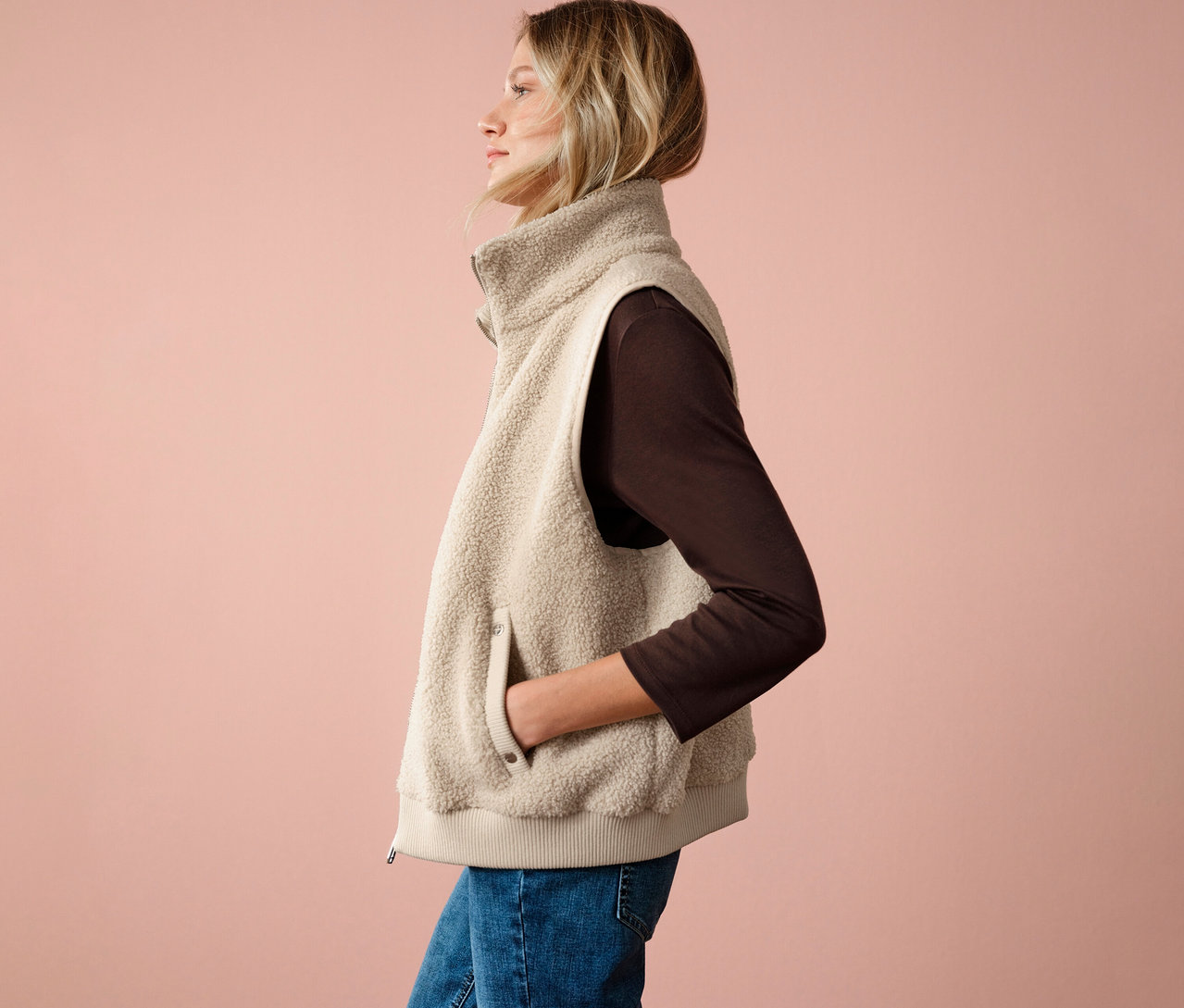 Žena v plyšové vestě Street One, hnědém topu a džínách, boční pohled na růžovém pozadí.