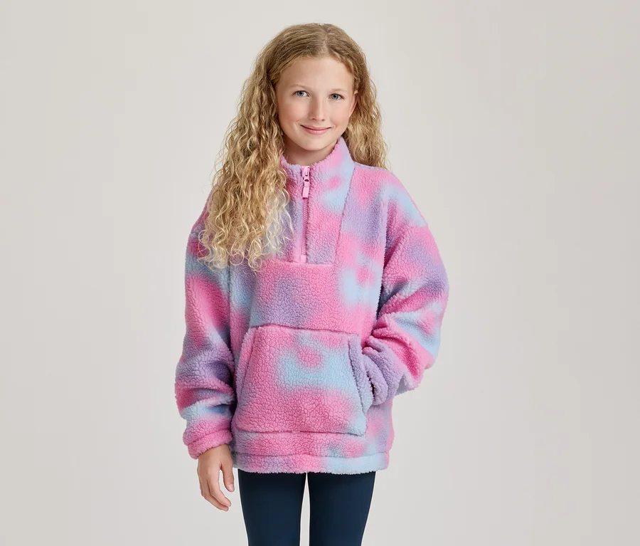Usmívající se dívka s kudrnatými blond vlasy v růžovo-fialovo-modrém tie-dye fleecovém pulovru s polovičním zipem a kapsou.