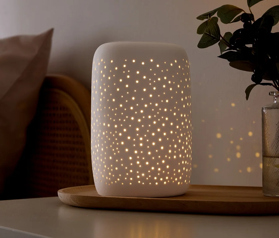 Bílá keramická lampa s malými perforacemi vyzařuje teplé světlo a vrhá hvězdné vzory na stěnu. Stojí na dřevěném podnosu na šedém povrchu, vedle béžového polštáře, proutěného nábytku a zelené rostliny ve skleněné váze.