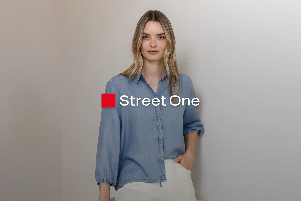 Blondýnka ve světle modré džínové košili a světlých kalhotách, stojí s rukou v kapse u šedé zdi. Logo Street One.