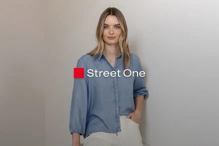 Blondýnka ve světle modré džínové košili a světlých kalhotách, stojí s rukou v kapse u šedé zdi. Logo Street One.