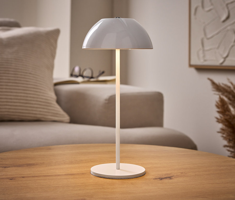 Osvětlená bílá akumulátorová stolní lampa s LED na dřevěném stole, s béžovou pohovkou, knihou a abstraktním uměním v pozadí.